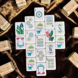 Puede incluir: Una colecci&oacute;n de fichas de mahjong rectangulares blancas con ilustraciones coloridas de paraguas, monos, flores y marcos de bamb&uacute;. Varias fichas est&aacute;n marcadas con los n&uacute;meros del 1 al 9, las letras N y W, y la palabra JOKER.