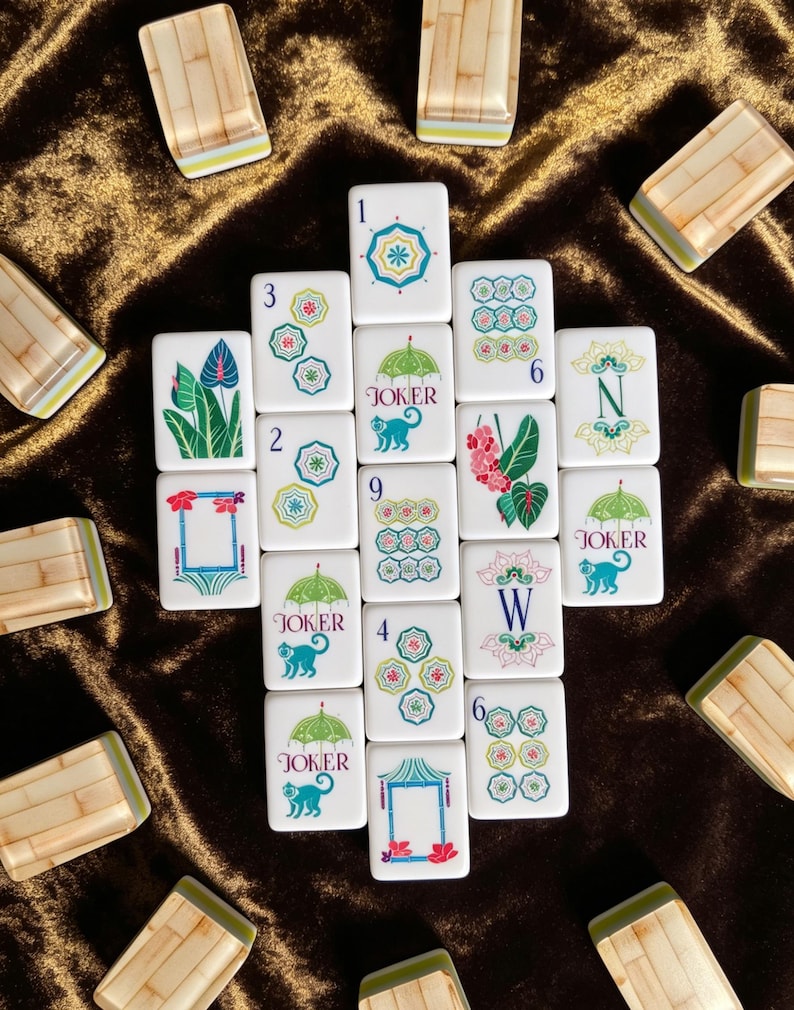 Puede incluir: Una colecci&oacute;n de fichas de mahjong rectangulares blancas con ilustraciones coloridas de paraguas, monos, flores y marcos de bamb&uacute;. Varias fichas est&aacute;n marcadas con los n&uacute;meros del 1 al 9, las letras N y W, y la palabra JOKER.