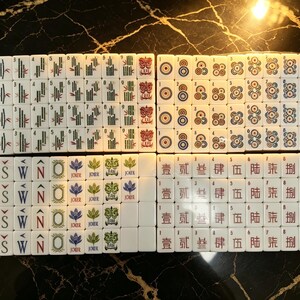May include: A complete set of white Mahjong tiles arranged in a grid. The tiles feature various colorful engraved designs including bamboo sticks, circular patterns, directional letters E, S, W, N, Joker symbols, and Chinese characters with red lotus icons.