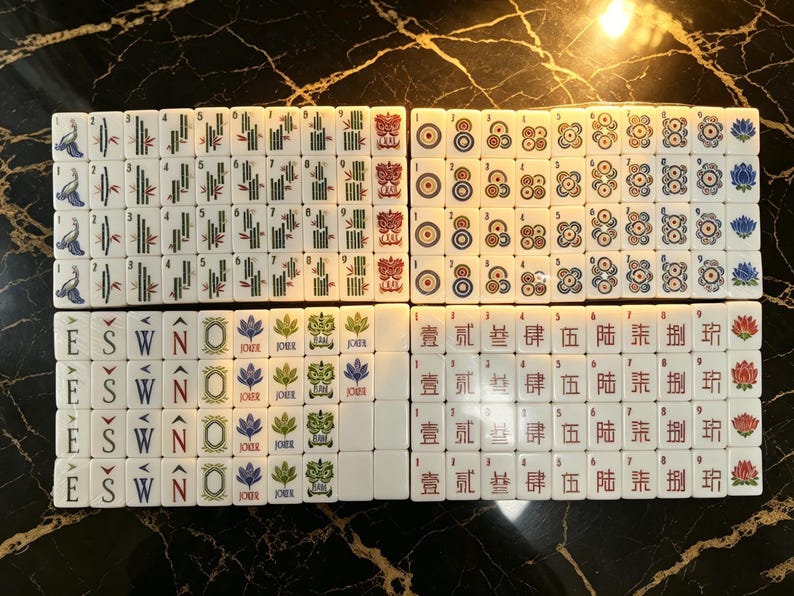 May include: A complete set of white Mahjong tiles arranged in a grid. The tiles feature various colorful engraved designs including bamboo sticks, circular patterns, directional letters E, S, W, N, Joker symbols, and Chinese characters with red lotus icons.