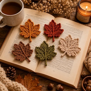 May include: Five crocheted maple leaf appliques in shades of mustard yellow, deep red, brown, olive green, and cream. These textured fabric leaves are arranged on the open pages of a book, featuring visible stitching and detailed leaf veins.