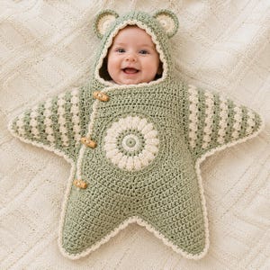 May include: A star-shaped crochet baby bunting in sage green and cream. The garment features a hood with bear ears, a central circular floral motif, and three wooden toggle buttons along the side. The sleeves and body are textured with a striped pattern.