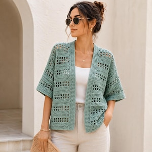 Puede incluir: Un cárdigan de ganchillo color verde salvia abierto por delante, con un patrón de rejilla geométrica, mangas cortas y dobladillo recto. La prenda se lleva sobre una camiseta blanca sencilla y pantalones claros, mostrando un diseño de punto ligero.