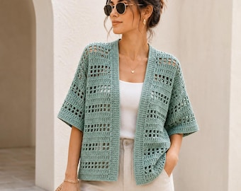 May include: A sage green open-front crochet cardigan featuring a geometric grid pattern, short sleeves, and a straight hemline. The garment is worn over a simple white tank top and light-coloured trousers, showcasing a textured, lightweight knit design.
