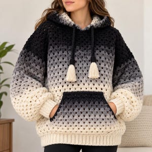 Könnte beinhalten: Ein langärmeliger Häkel-Kapuzenpullover mit einem Farbverlauf, der von Schwarz oben über Grau in der Mitte bis zu Creme unten reicht. Das Kleidungsstück verfügt über eine Kängurutasche vorne, eine Kapuze mit schwarzen Kordelzügen und cremefarbene Quasten.
