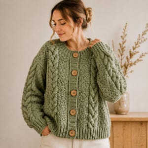 May include: A sage green long-sleeve cardigan featuring a chunky cable knit pattern on the sleeves and a textured knit body. The garment has a round neckline, a ribbed hem and cuffs, and is fastened down the front with five round wooden buttons.