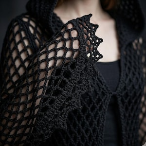 Könnte beinhalten: Eine schwarze Kapuzenjacke mit einem durchbrochenen Häkelnetzmuster. Das Kleidungsstück ist in einem rautenförmigen Netzdesign gefertigt und verfügt über einen dekorativen, spitz zulaufenden Bogenrand an den Ärmelbündchen und der Vorderseite.