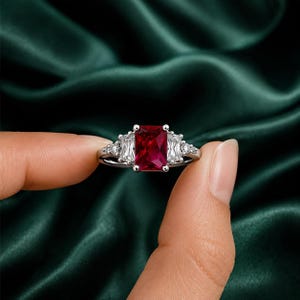 Puede incluir: Un anillo de tono plateado con una gran piedra central rectangular de color rojo intenso, flanqueada por dos piedras laterales rectangulares transparentes y dos diminutas piedras redondas transparentes en una banda de metal pulido.