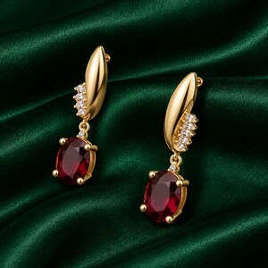 May include: A pair of gold drop earrings featuring a polished gold leaf-shaped top accented with a row of small clear stones. Each earring suspends a large, oval-cut deep red gemstone held in a four-prong setting, creating a dangling design.