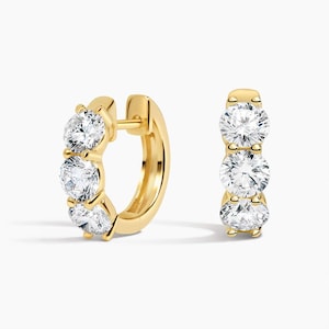 May include: A pair of gold hoop earrings, each featuring three round, clear, faceted stones set in a vertical row along the front of the hoop. The earrings have a polished gold finish and a hinged closure mechanism for secure wear.