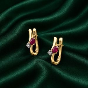 May include: A pair of gold hoop earrings featuring a polished yellow gold frame, each set with one oval-cut deep red ruby and one small round-cut clear white diamond, designed with a secure hinged latch closure mechanism.