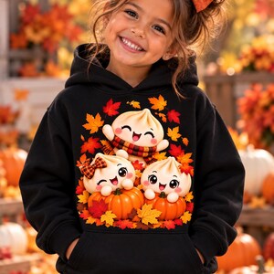 May include: A black hooded sweatshirt featuring a graphic print of three smiling, cartoon-style steamed buns. Two buns sit inside orange pumpkins, while one sits above them. The design is surrounded by vibrant red, orange, and yellow autumn leaves.