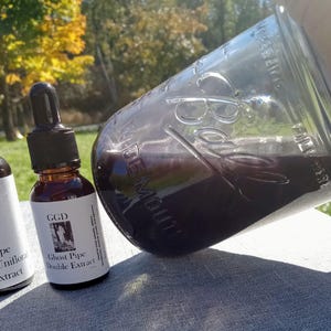 May include: A clear glass jar and a small amber glass bottle with a dropper. The jar is filled with a dark purple liquid. The bottle has a label that reads "GGD Ghost Pipe Double Extract". The background is a blurred view of trees.