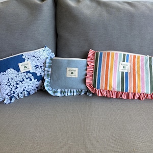 Peut inclure: Trois pochettes zippées avec des bords à volants et une étiquette rectangulaire blanche portant l'inscription Atelier. La pochette de gauche est bleue à motif floral blanc, celle du centre est bleu clair uni avec un volant à carreaux verts et celle de droite est rayée.