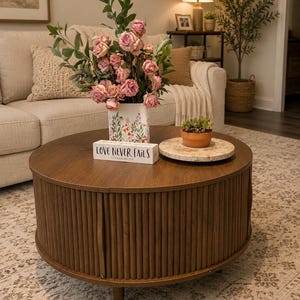 Puede incluir: Una mesa de centro redonda de madera marrón oscuro con una base de listones estilo tambor y patas cortas. Encima hay un jarrón blanco con estampado floral que contiene rosas rosadas y vegetación, una pequeña suculenta y un letrero de bloque.