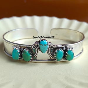 May include: A silver cuff bracelet featuring five teardrop-shaped turquoise stones set in metal bezels. The centre stone is elevated above the others, flanked by two stones on each side. The text JewelsSilverArt92 is printed above the centre stone.