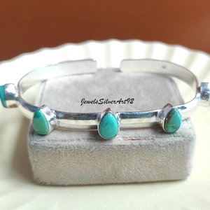 May include: A silver cuff bracelet featuring five teardrop-shaped turquoise stones set along the band. The metal has a polished finish, and the text JewelsSilverArt92 is printed in black on the surface of the bracelet. The design is open-ended.