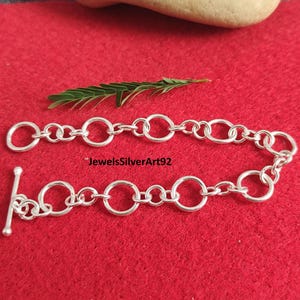 Op de afbeelding: Een zilveren kettingarmband met een reeks onderling verbonden cirkelvormige schakels en een staafsluiting. De tekst JewelsSilverArt92 is in het zwart gedrukt nabij het midden van de armband, die bestaat uit gepolijste zilverkleurige ringen.