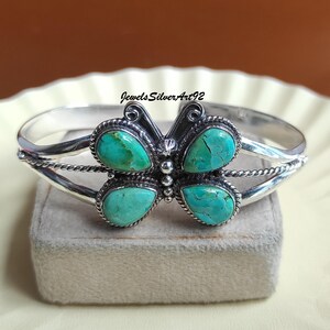 Könnte beinhalten: Ein silbernes Armband mit einem zentralen Schmetterlingsmotiv, das mit vier birnenförmigen Türkissteinen verziert ist. Die Metallarbeit umfasst gedrehte Drahtdetails und kleine Silberperlen. Der Text JewelsSilverArt92 ist über dem Armband sichtbar.