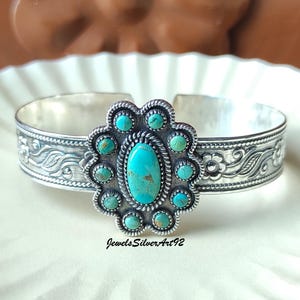 May include: A silver cuff bracelet featuring a central oval turquoise stone surrounded by ten smaller round turquoise stones in a floral arrangement. The silver band is decorated with embossed floral and leaf patterns and the text JewelsSilverArt92.