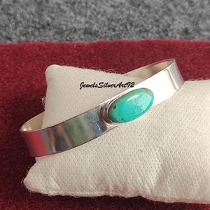 May include: A polished silver cuff bracelet featuring a single, smooth, oval-shaped turquoise stone set in the centre. The metal band has a reflective surface and the text JewelsSilverArt92 is printed in black ink above the turquoise stone.