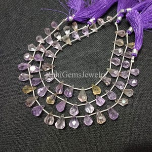 May include: A strand of faceted, pear-shaped amethyst and citrine gemstones arranged in a curved pattern, accented with vibrant purple tassels at the top. The stones display translucent shades of light purple and pale yellow, strung on thin wire.