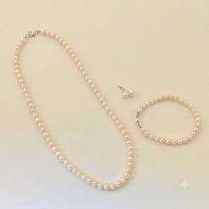 Può includere: Un set di gioielli composto da una collana lunga, un braccialetto e un paio di orecchini a lobo, tutti caratterizzati da perle rotonde e uniformi di colore rosa chiaro con una finitura liscia e lucente e chiusure in metallo argentato.
