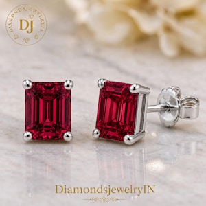 May include: A pair of stud earrings featuring emerald-cut red gemstones set in polished silver-toned metal. Each earring has four prongs securing the rectangular stone, with a standard post and butterfly back closure visible on one of the earrings.