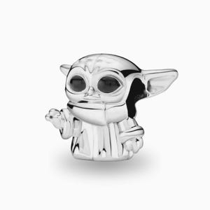 May include: A polished silver charm shaped like a small creature with large, dark, oval eyes, pointed ears, and a robe with vertical folds. The figure is depicted with one hand raised in a gesture, featuring a smooth, reflective metallic finish.