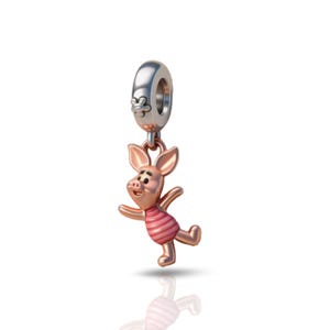 May include: A rose gold-toned metal charm featuring a small pig character with large ears and a striped shirt. The charm is attached to a silver-toned cylindrical bead with a small flower detail on the side, designed for a bracelet or necklace.