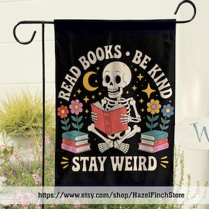 Könnte beinhalten: Eine schwarze Gartenflagge mit einem Skelett, das im Schneidersitz ein rotes Buch liest. Der Text lautet READ BOOKS BE KIND STAY WEIRD. Das Design zeigt einen gelben Halbmond, Sterne, bunte Blumen und mehrere Bücherstapel.
