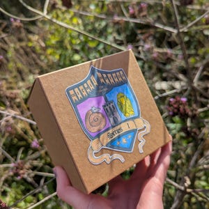 May include: A square brown cardboard box featuring a shield-shaped sticker with the text "Micah Morphs The Collection Series 1". The sticker displays a coiled creature, a stone tower, and a yellow bird illustration on a blue and purple background.