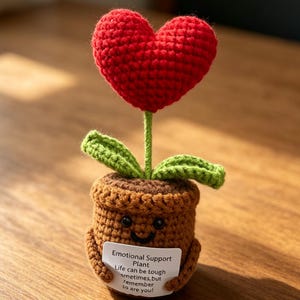 May include: A crocheted plant in a brown pot with a red heart-shaped flower and two green leaves. The pot has a smiling face and holds a white sign that reads: Emotional Support Plant, Life can be tough sometimes, but remember so are you!