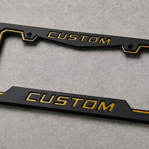 May include: A rectangular matte black license plate frame featuring a gold-colored inner border and the word CUSTOM embossed in gold lettering on both the top and bottom sections of the frame, including four pre-drilled mounting holes.