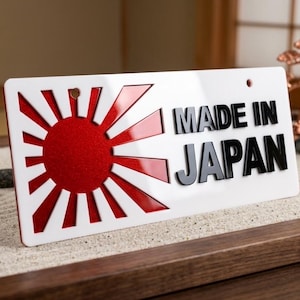 May include: A rectangular white sign featuring a red sunburst graphic on the left side and the black text MADE IN JAPAN on the right. The sign has two small circular holes at the top and a glossy finish, presenting a clean and high-contrast design.