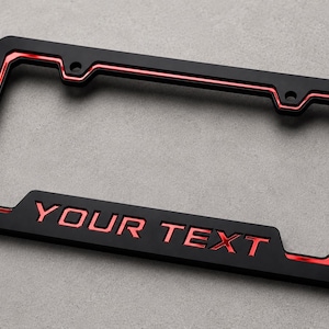 May include: A rectangular license plate frame featuring a matte black finish with metallic red accents along the inner border and the words YOUR TEXT embossed in red lettering at the bottom center, designed with four mounting holes for attachment.