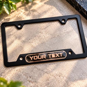 May include: A rectangular black license plate frame featuring a centered horizontal bar at the bottom with a copper-colored border and the text YOUR TEXT embossed in matching copper lettering against a black background.