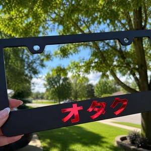 Pode incluir: Uma moldura de matrícula retangular preta mate com o texto japonês em katakana "オタク" num acabamento vermelho brilhante e refletor. A moldura inclui dois orifícios de montagem no topo e uma borda inferior larga.