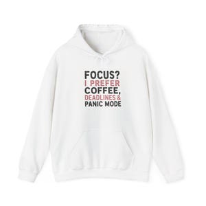 Könnte beinhalten: Ein weißer Kapuzenpullover mit einer Kängurutasche auf der Vorderseite und langen Ärmeln. In der Mitte der Brust befindet sich der schwarze und rosafarbene Text: FOCUS? I PREFER COFFEE, DEADLINES & PANIC MODE. Der Hoodie hat weiße Kordelzüge.