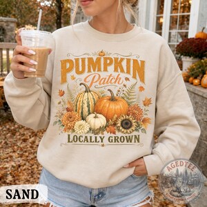 May include: A sand-colored crewneck sweatshirt featuring a graphic print with the text PUMPKIN PATCH LOCALLY GROWN. The design includes illustrations of orange and green pumpkins, white gourds, yellow sunflowers, orange chrysanthemums, and autumn leaves.