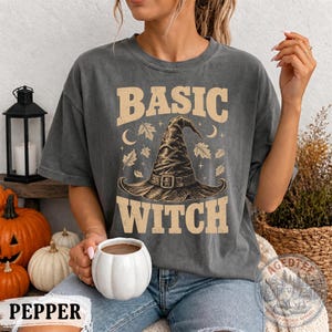 May include: A charcoal gray short-sleeve t-shirt featuring a graphic of a pointed witch hat surrounded by autumn leaves, stars, and crescent moons. The text BASIC WITCH is printed in a distressed beige font above and below the central hat illustration.