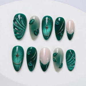 Peut inclure: Un ensemble de dix faux ongles en forme d'amande dans des tons vert émeraude et crème. Les motifs incluent des finitions magnétiques œil-de-chat, des feuilles en relief, des étoiles dorées, des lignes fines et des gouttes d'eau 3D brillantes.