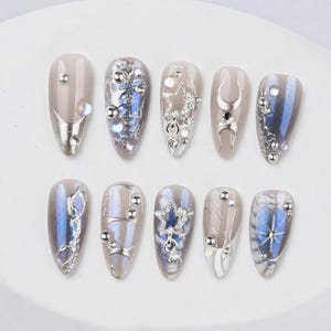 Peut inclure: Un ensemble de dix faux ongles en forme d'amande avec une base nude, des accents bleu métallisé, des détails chromés argentés et des ornements en relief incluant des étoiles, des lunes, des sphères et des strass disposés en motifs.