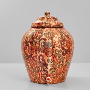 May include: A copper water dispenser with a rounded body and a matching lid. The surface is covered in a detailed, multi-colored floral and paisley pattern in shades of orange, red, green, and black. A small red circular tap is at the base.