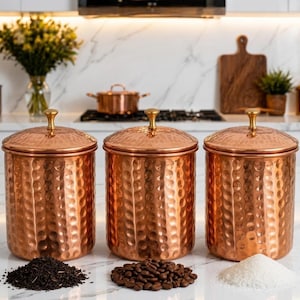 May include: A set of three cylindrical copper canisters with a hammered texture and polished gold-tone knobs on the lids. Each canister is filled with a different ingredient: loose tea leaves, roasted coffee beans, and white granulated sugar.
