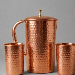 Op de afbeelding: Een gehamerde koperen kan met bijpassend deksel en twee cilindrische gehamerde koperen bekers. De kan heeft een gebogen handvat en een kleine knop op het deksel. De set heeft een metallic afwerking met een duidelijk textuurpatroon.
