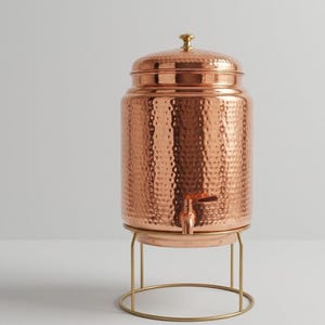 May include: A cylindrical copper beverage dispenser with a hammered texture, a matching lid with a small knob, and a metal spigot. The dispenser rests on a thin, circular gold-toned metal stand with four vertical legs and a base ring.