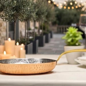 May include: A round, shallow frying pan with a hammered copper exterior and a polished silver-toned interior. The pan features a long, slender, gold-toned metal handle extending from the side, attached with a visible rivet near the rim.