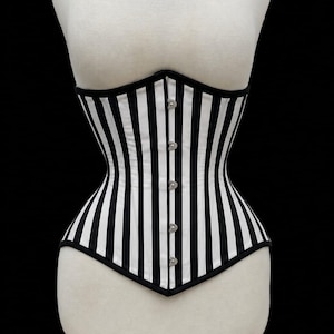 May include: A corset featuring a vertical black and white striped pattern. The garment has a straight neckline, a pointed hemline at the center front, and a front closure consisting of five silver-toned metal busk clasps with black trim edges.