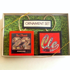 May include: A set of two Christmas ornaments in a box. One ornament is red with the number 22 and the text "The Streak" on it. The other ornament is red with the word "Cle" in a stylized font. The box has a green background and the text "ORNAMENT SET" on it.